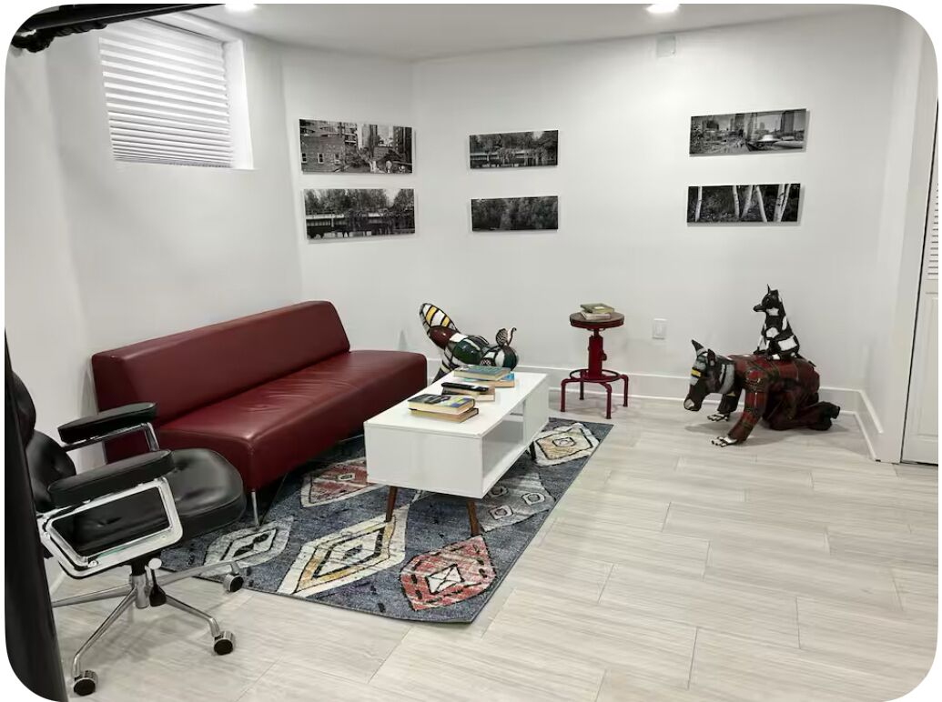 Ein modernes Wohnzimmer mit einem roten Sofa, einem weißen Couchtisch und einem gemusterten Teppich. An der Wand befinden sich Schwarz-Weiß-Fotos und auf einem roten Hocker eine kleine Statue.