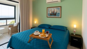 Habitació d'hotel amb un llit gran, una bandeja de petitdejuni amb croissants i suc d'aranja, un quadre d'un vaixell a la paret i vista al mar per la finestra.