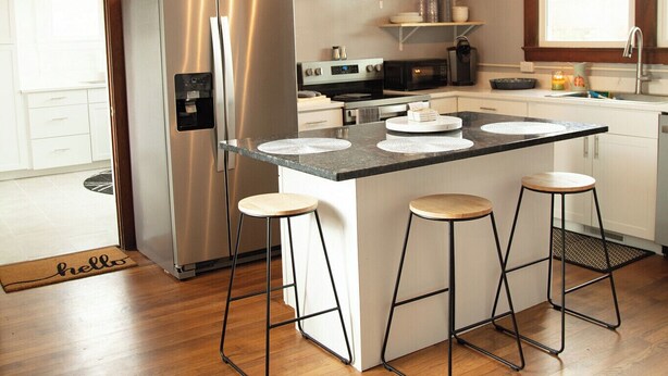 Uma cozinha moderna com geladeira de aço inoxidável, uma ilha central com banquetas, piso de madeira e luminária pendente.