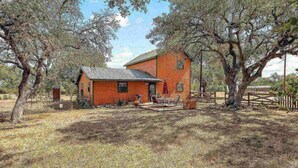 A rustic brick house with a small porch, surrounded by trees and a fenced yard. - NEW Artist's Retreat on 11 Acres, Pet-Friendly, Dark Skies, Nature Escape, Fire Pit, Games, Grill (New Braunfels)