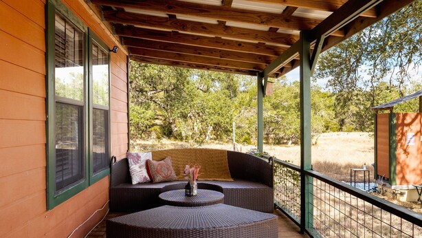 A covered outdoor seating area with wicker furniture and a small table, overlooking a natural landscape. - NEW Artist's Retreat on 11 Acres, Pet-Friendly, Dark Skies, Nature Escape, Fire Pit, Games, Grill (New Braunfels)
