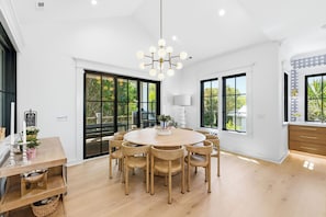 A spacious dining area with a round wooden table and chairs, large windows, and a modern chandelier. - LUXURY! 6 BR Sky Watch Views 2 Min Walk to E. Beach Shoals Club - 4 day min stay (Bald Head Island)