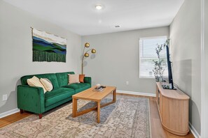 A living room with a green sofa, a wooden coffee table, a patterned rug, a wall hanging, a floor lamp, and a TV stand with a plant. - Family Fun w/Game Rm, WFH Office: 8 Min to Beach (Ocean Isle Beach)