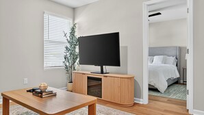 A modern living room with a wooden TV stand, a flat-screen TV, a dining table, and a bed in the adjacent room. - Family Fun w/Game Rm, WFH Office: 8 Min to Beach (Ocean Isle Beach)