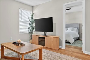 A modern living room with a wooden TV stand, a flat-screen TV, a dining table, and a bed in the adjacent room. - Family Fun w/Game Rm, WFH Office: 8 Min to Beach (Ocean Isle Beach)