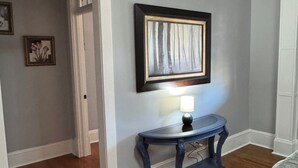 A hallway with a wooden floor, a dark blue console table with a lamp, and a framed picture on the wall. - The Wisteria House invites you to relax and enjoy! (Shelby)