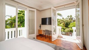 A bedroom with a large bed, a desk, and a TV. There are sliding glass doors leading to a balcony with a view of greenery. - Palais Elya (Rabat)