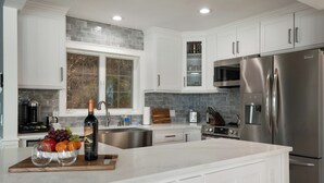 Une cuisine moderne avec des meubles blancs, un réfrigérateur en acier inoxydable et une planche à découper en bois sur laquelle sont disposés des fruits et des verres à vin.