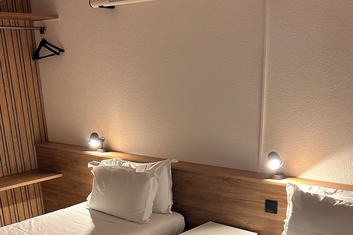 A hotel room with two beds, a wooden headboard, a nightstand, and a wall-mounted heater.