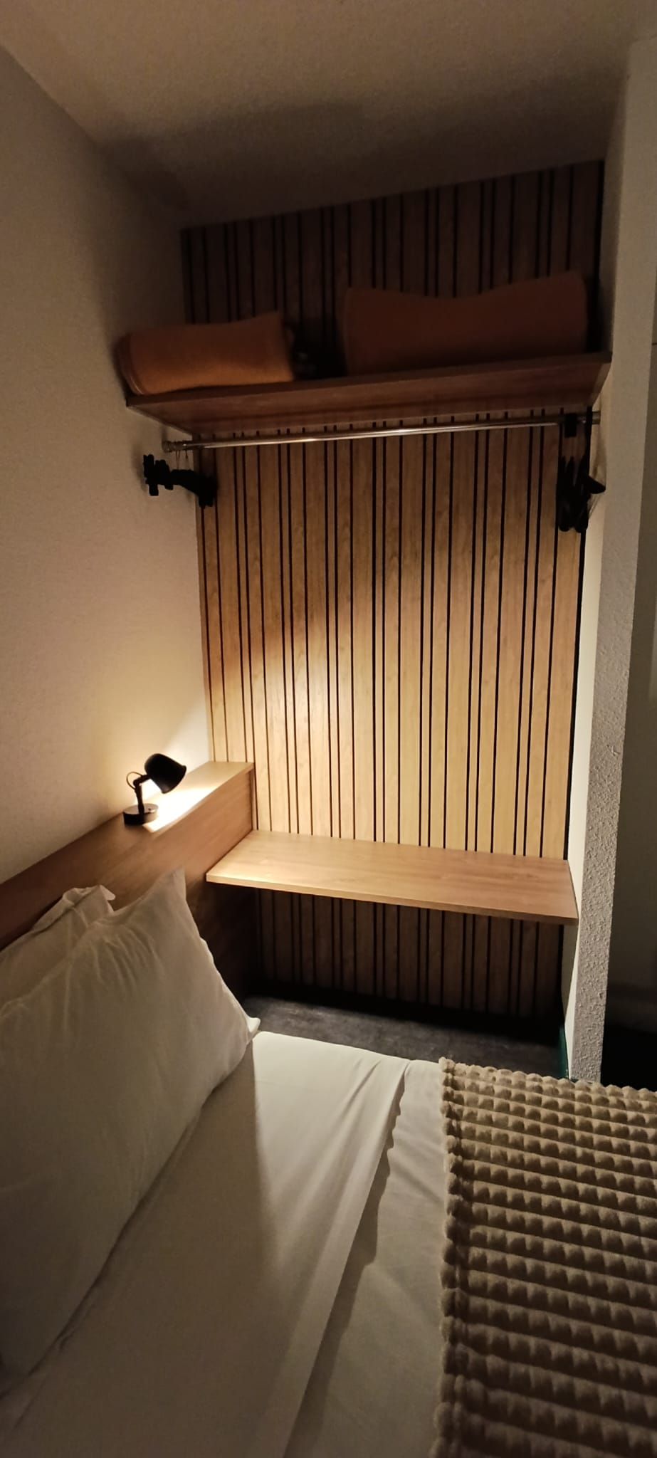 A compact hotel room with a bunk bed, a bedside table with a lamp, and a wooden slat wall panel.