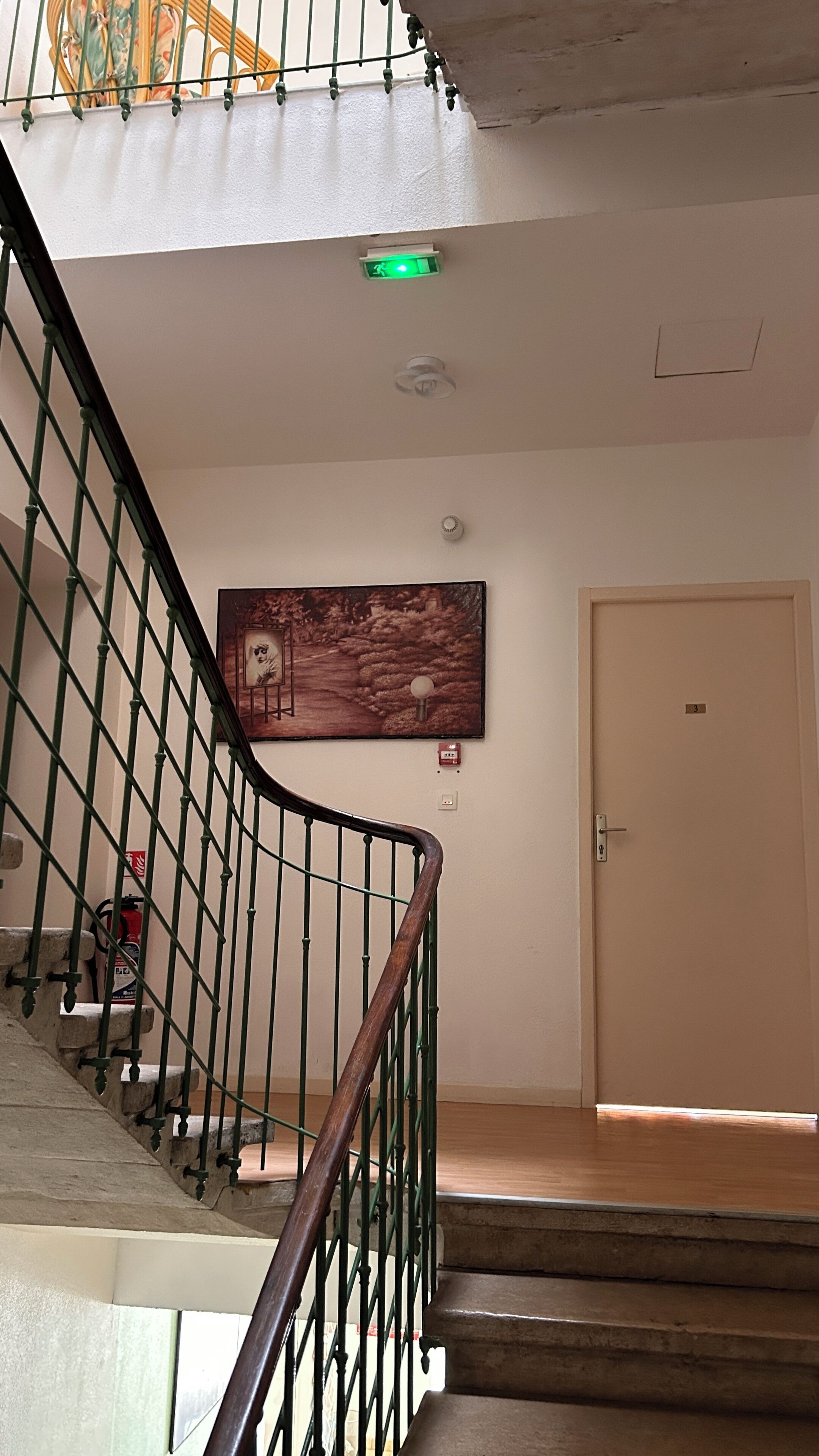 Una escalera con barandilla de metal, un extintor, un cuadro enmarcado, una puerta con número y una lámpara en el techo.