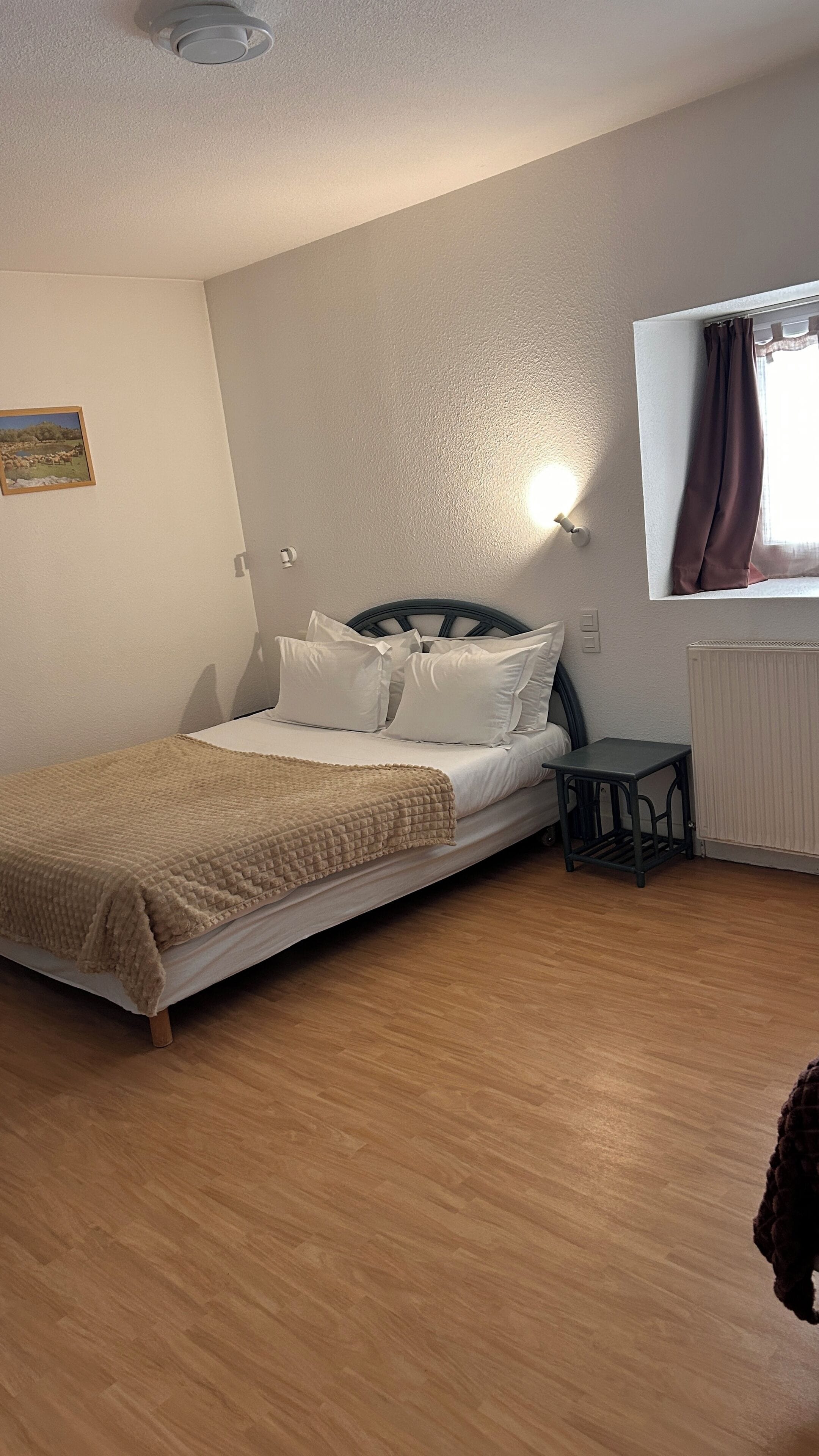 Un dormitorio con cama, una mesita, un radiador y una ventana con cortinas.