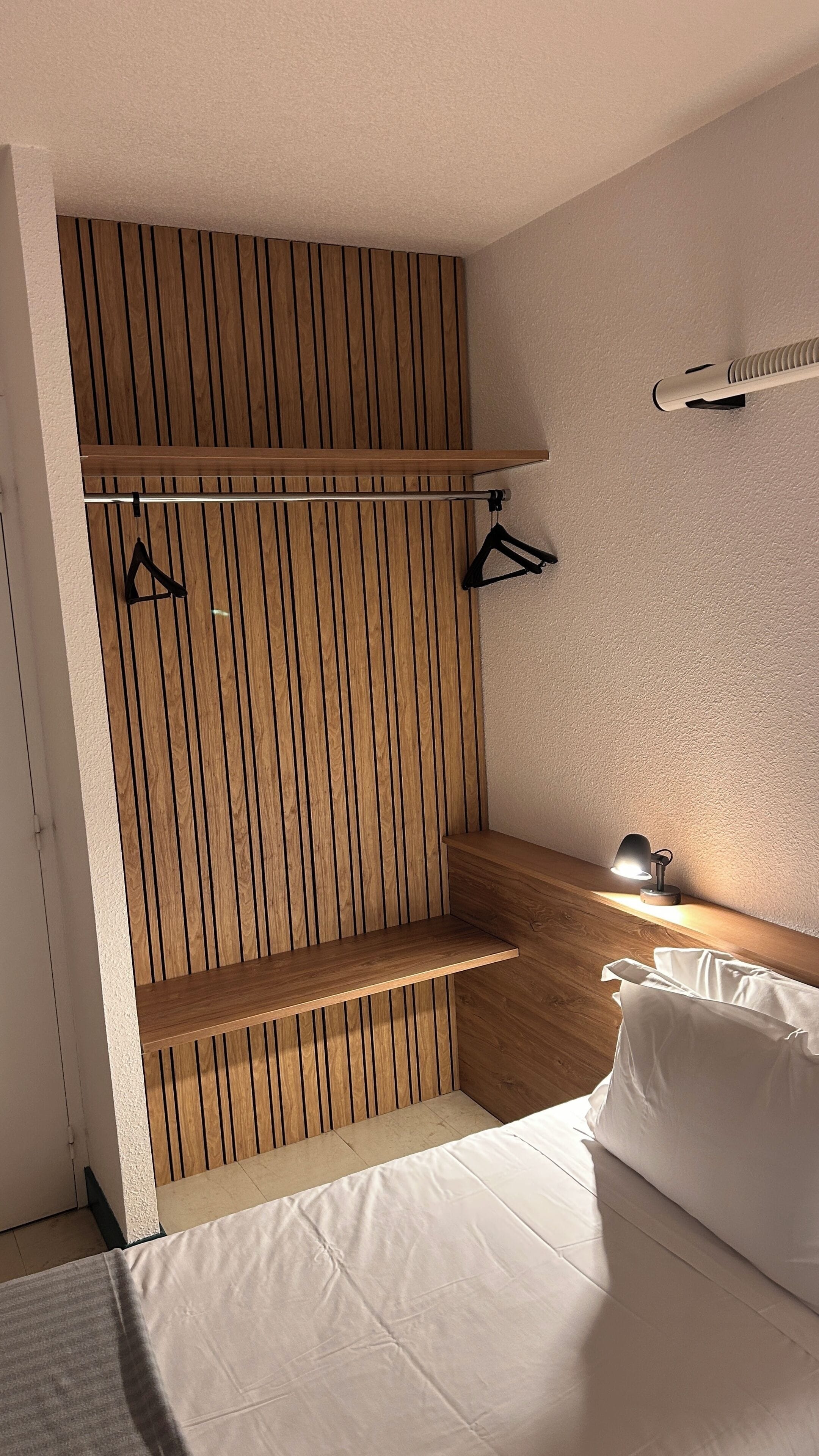 A bedroom with a wooden slat wall, a bed with white bedding, a bedside table with a lamp, and a hanging light fixture.