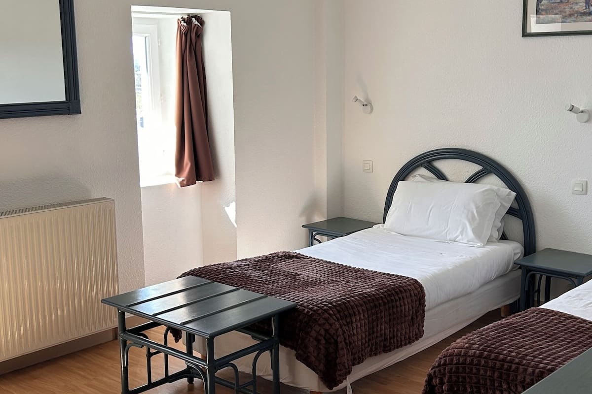 A room with a bed, a table, a chair, a radiator, a window with curtains, and a picture on the wall.