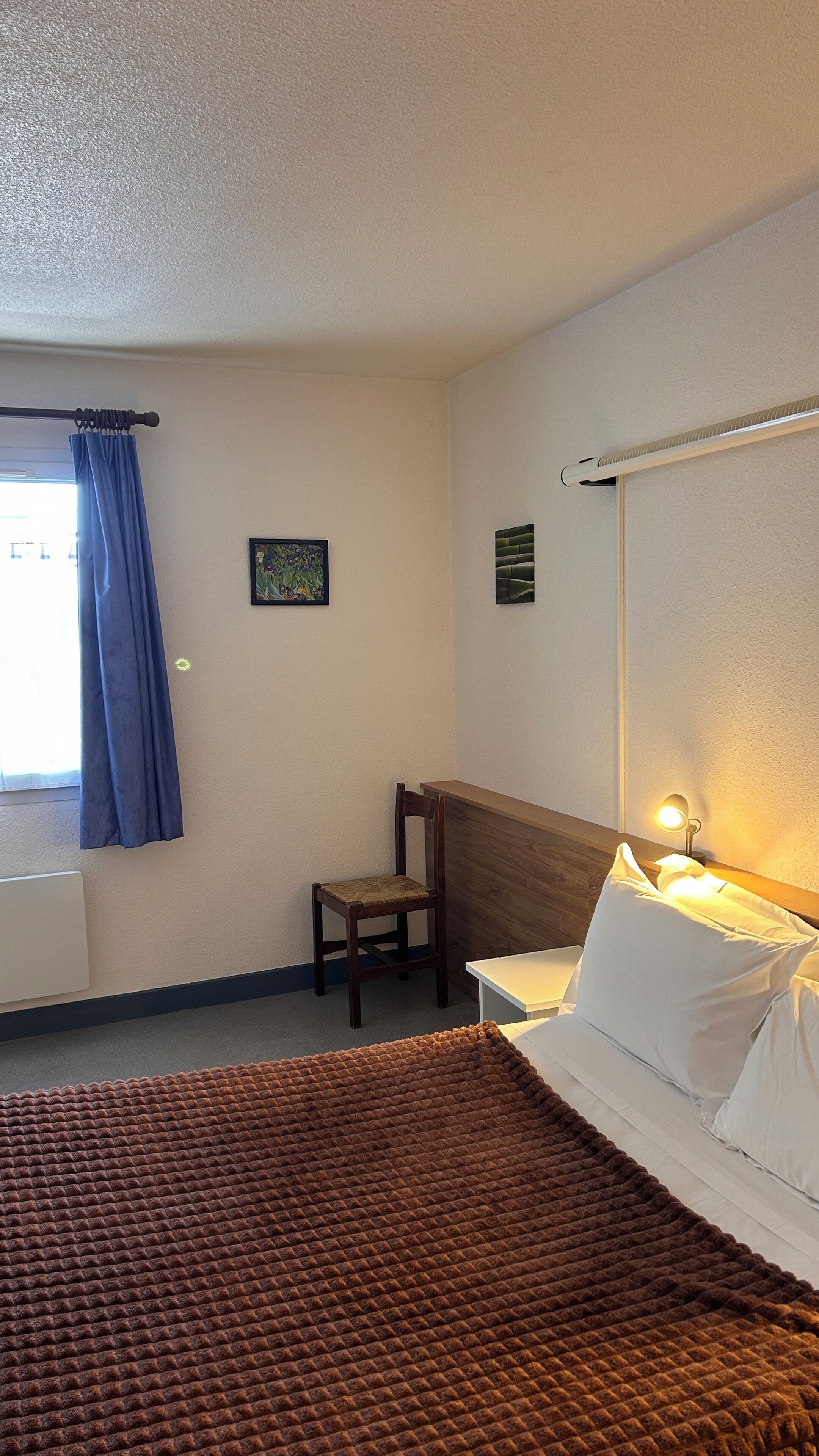 A hotel room with a bed, a chair, a desk, a window with curtains, and a wall-mounted lamp.