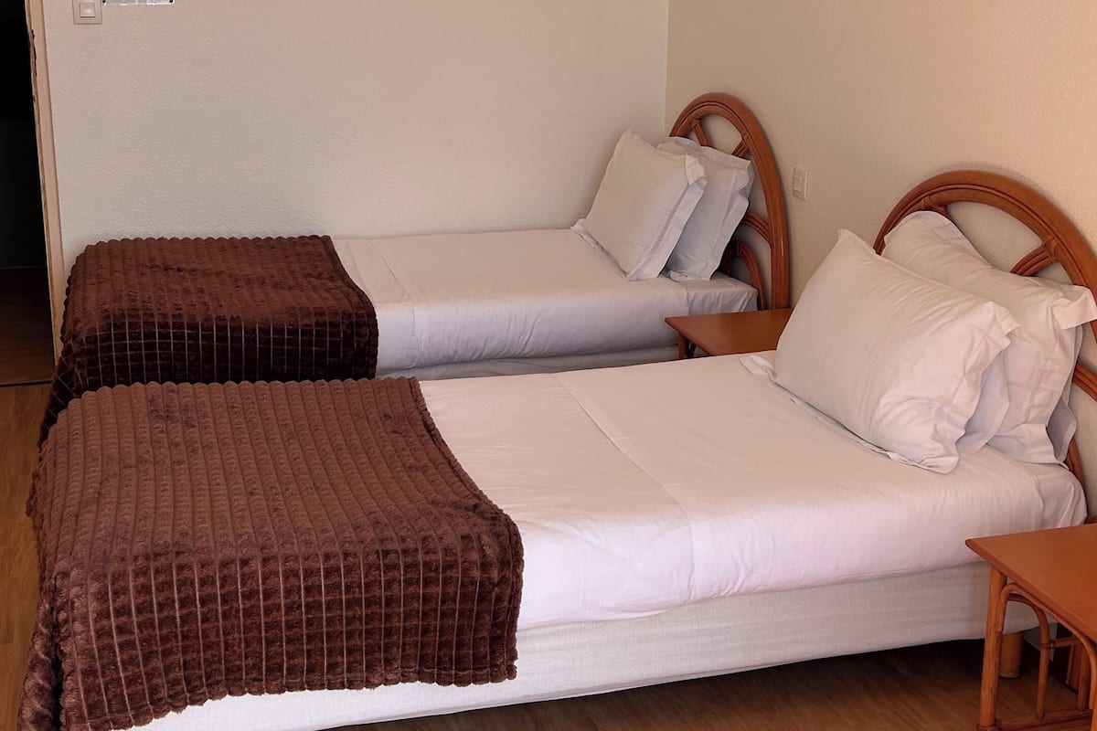 A room with two beds, a wooden floor, a small wooden table, and a wall-mounted light switch.