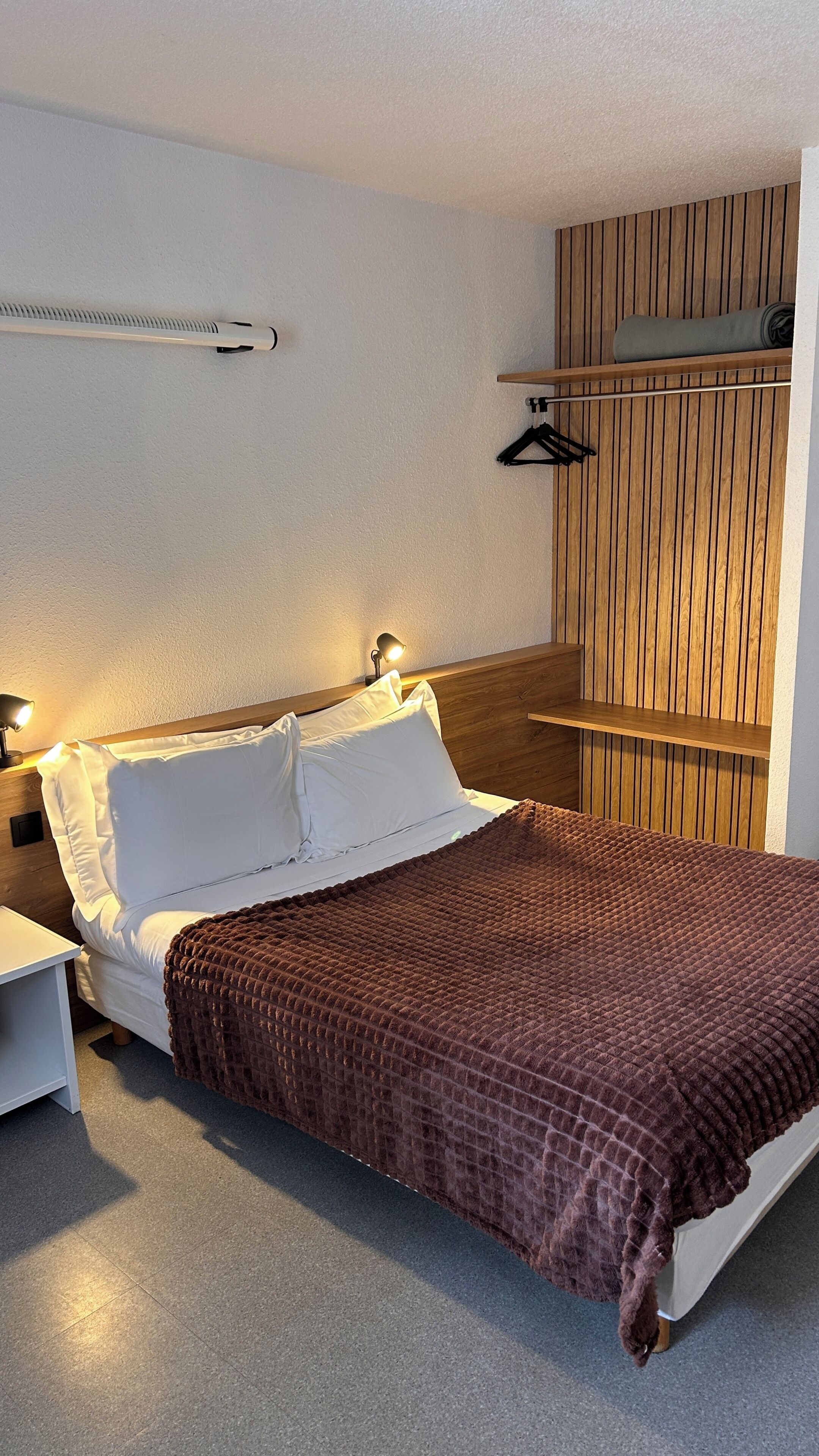 Ein ordentlich bezogenes Bett mit Kissen, einer Nachttischlampe, einem hölzernen Kopfteil, einem kleinen Regal mit einer zusammengefalteten Decke und einem an der Wand befestigten Kleiderhaken.