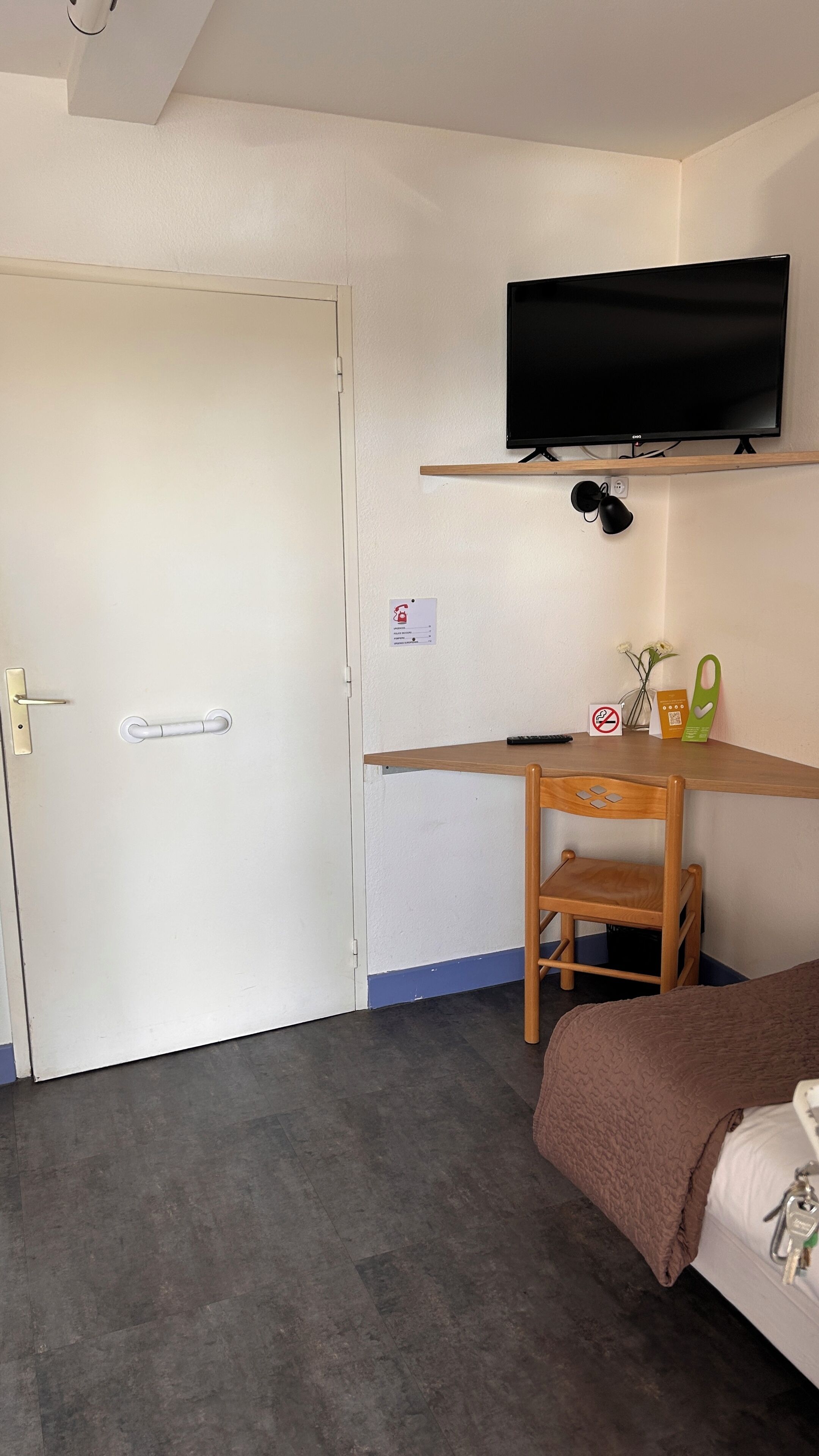 Un dormitorio con cama, un escritorio con silla, un televisor montado en la pared y una puerta con manija.