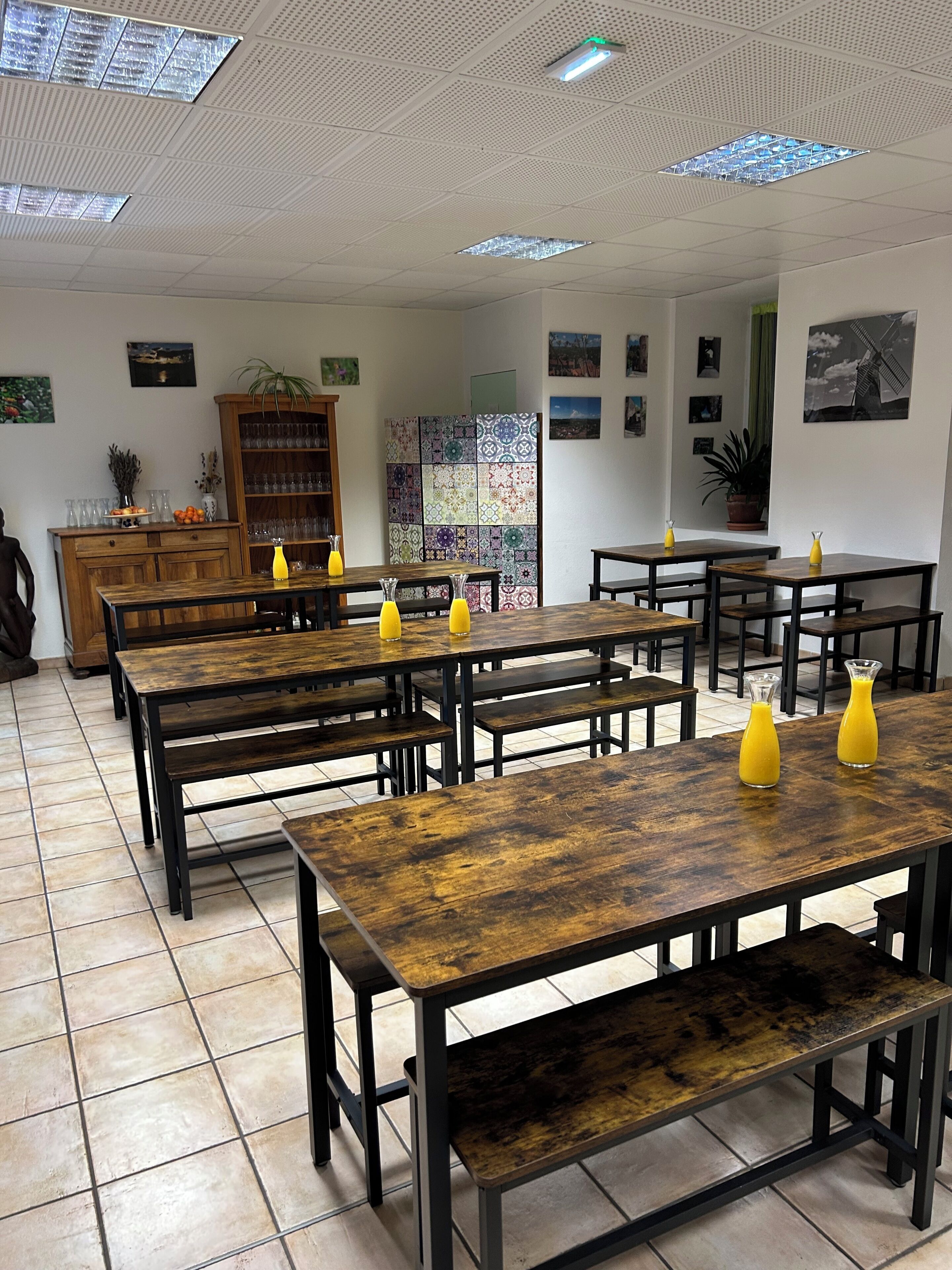 A dining area with wooden tables and benches, a tiled floor, and a ceiling with perforated panels and recessed lighting.