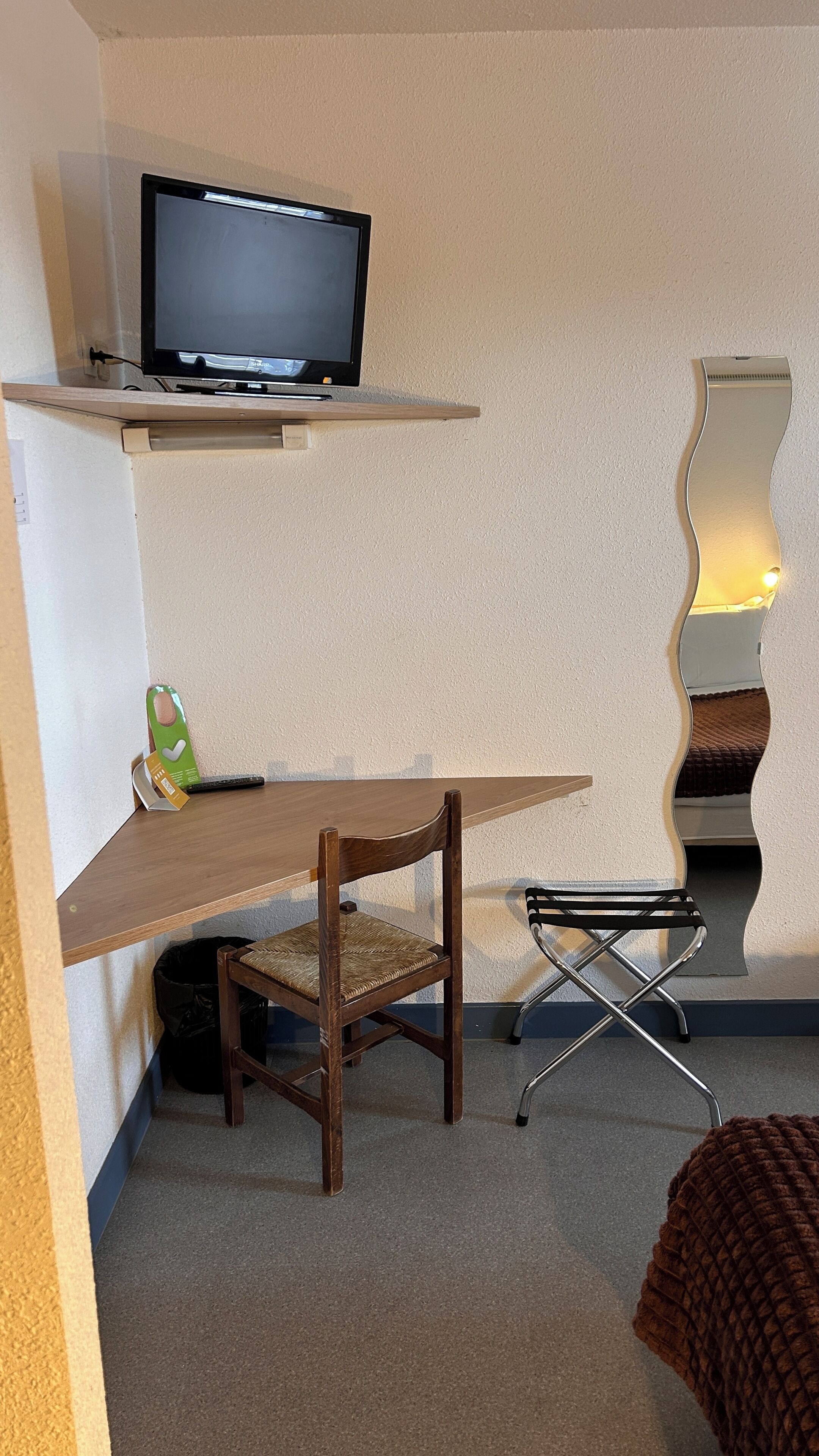 Une petite pièce avec un bureau en bois, une chaise, un téléviseur posé sur une étagère, une poubelle noire et un miroir doté d’un cadre ondulé.