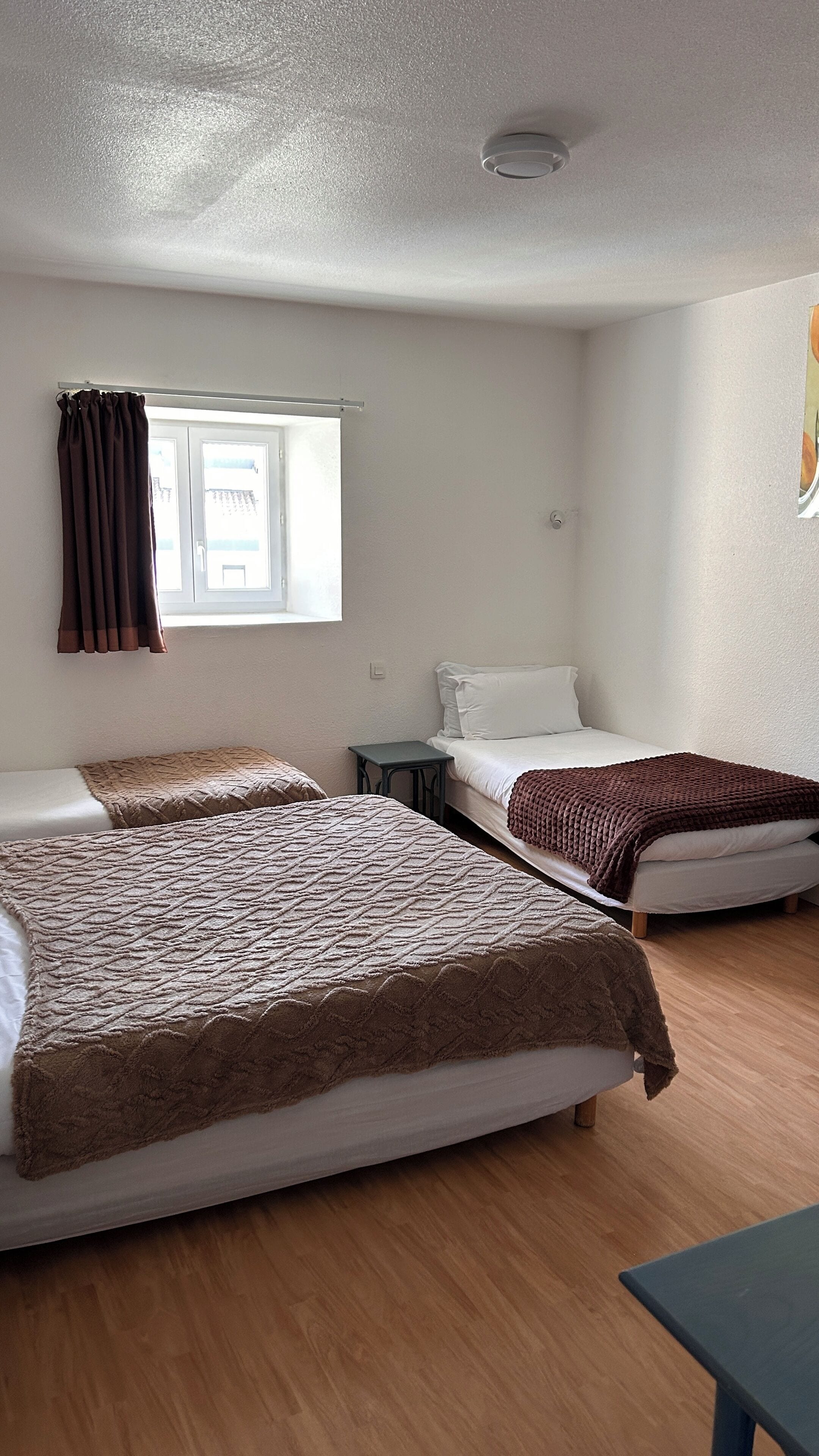 A room with two beds, a small table, a window with curtains, and a ceiling light.
