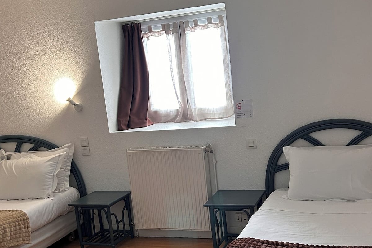 A bedroom with two beds, a window with curtains, a radiator, and a small table.