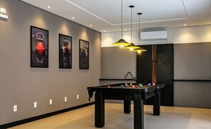 A modern indoor pool table with a pool cue, surrounded by a neutral-toned room with framed artwork on the walls. - Estadia em prédio com piscina a poucos passos da praia em Florianópolis - STIs (Florianópolis)