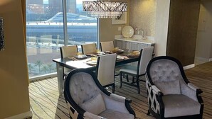 A hotel room with a dining area, two armchairs, a sofa, and a view of the cityscape.