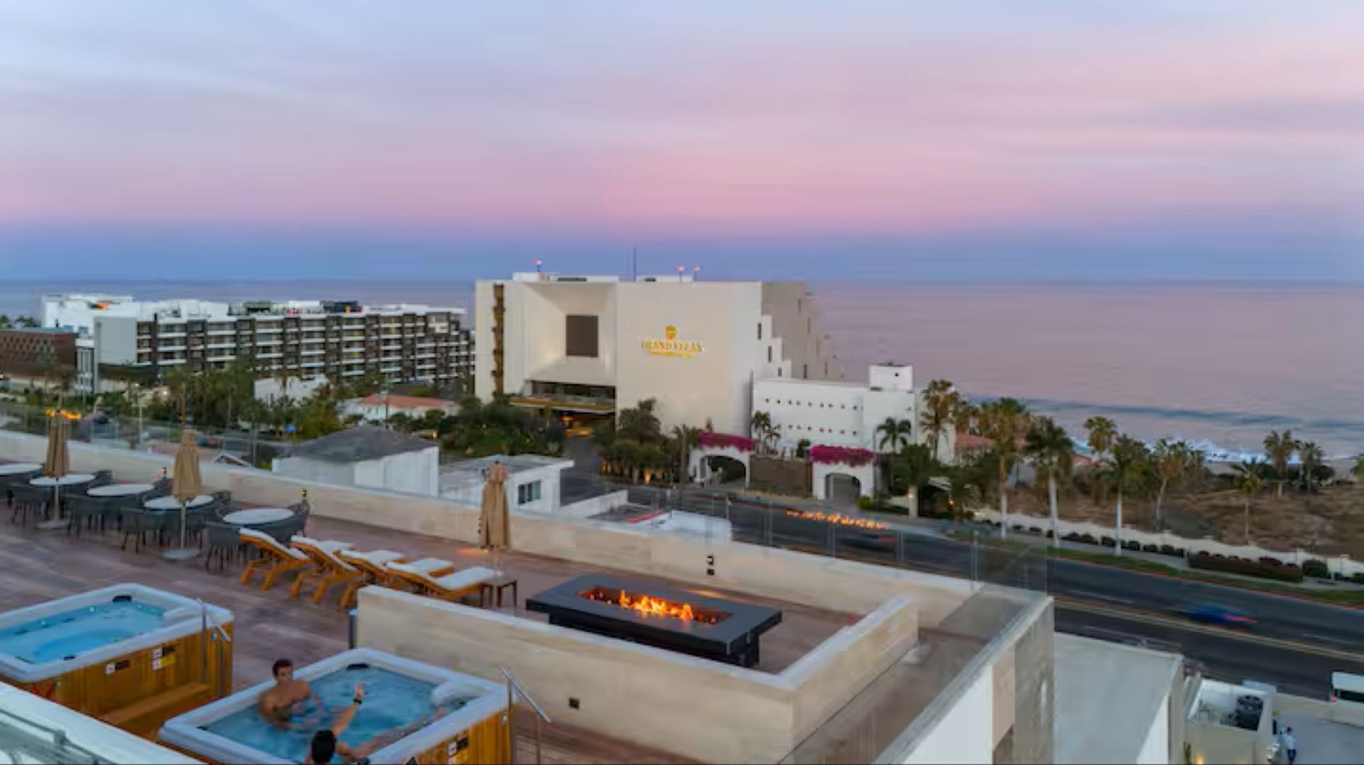 Un espace piscine aménagé sur le toit, avec des chaises longues et un foyer, offrant une vue sur la plage et l’océan au coucher du soleil.
