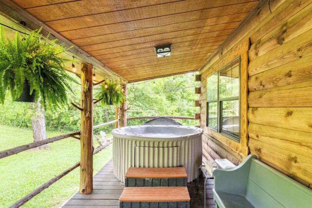 A wooden cabin with a hot tub on a wooden deck, surrounded by greenery.