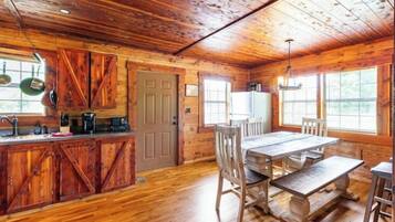 A rustic cabin interior with a wooden dining table, benches, and a kitchen area with wooden cabinets and a refrigerator.