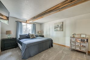 Quarto com cama, mesas de cabeceira, cômoda, janela com cortinas, um quadro na parede e teto com vigas aparentes.