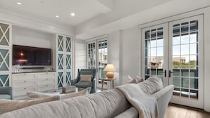 A spacious living room with a large grey sofa, a television, and a wooden coffee table. There are large windows with shutters and a view of the outside. - Charming Coastal Escape – Private Beach & Pool Access In Rosemary Beach (Rosemary Beach)