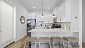A modern kitchen with white cabinetry, a large island with bar stools, stainless steel appliances, and pendant lighting. - By The Beach: 2BR House (Orange Beach)