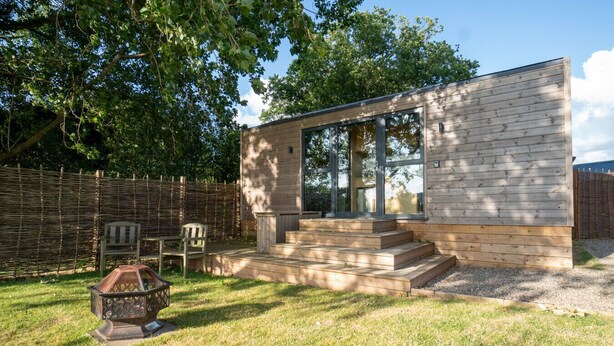 A modern, single-story wooden house with large windows, a wooden deck, and a garden area with a chair and a decorative lantern.