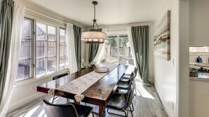 Uma sala de jantar com uma mesa de madeira comprida, cadeiras pretas, um candelabro e vista através de grandes janelas com cortinas.