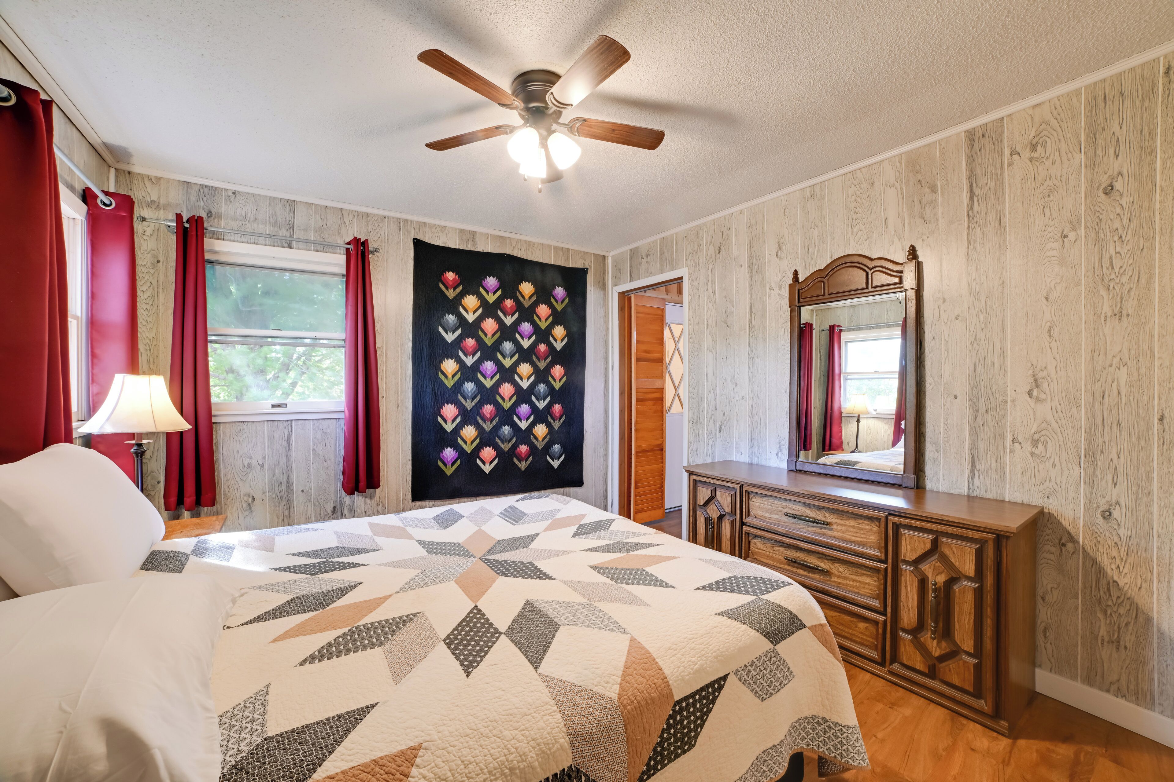 A bedroom with a bed, a dresser, a ceiling fan, a window with curtains, and a quilt on the wall.