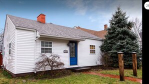 A white single-story house with a blue door, a chimney, and a small tree in front.