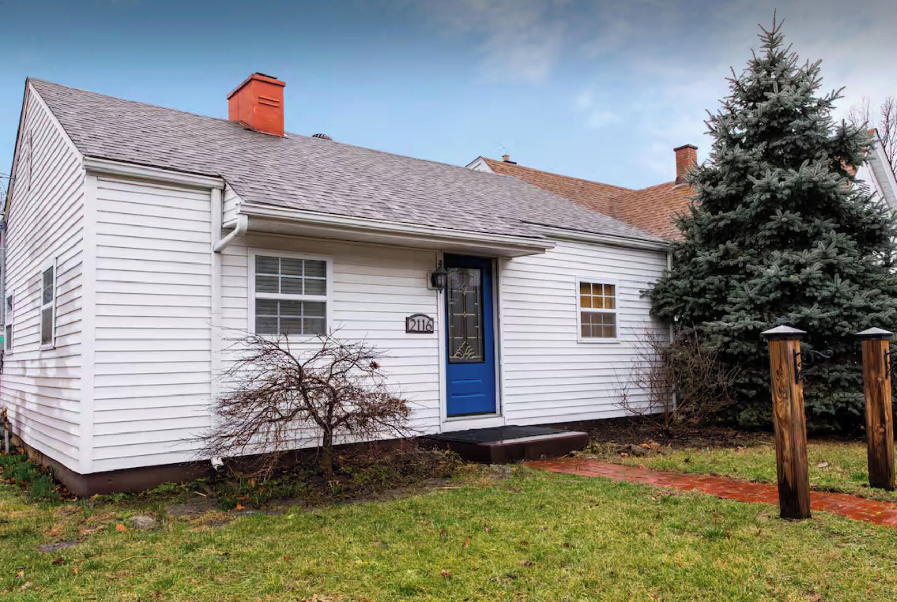 A white single-story house with a blue door, a chimney, and a small tree in front.