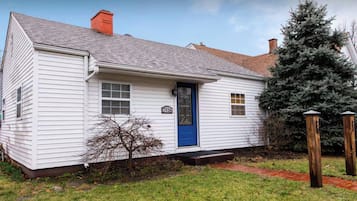 A white single-story house with a blue door, a chimney, and a small tree in front.