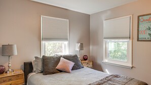Une chambre à coucher avec un lit, deux tables de chevet, des lampes et deux fenêtres équipées de stores.