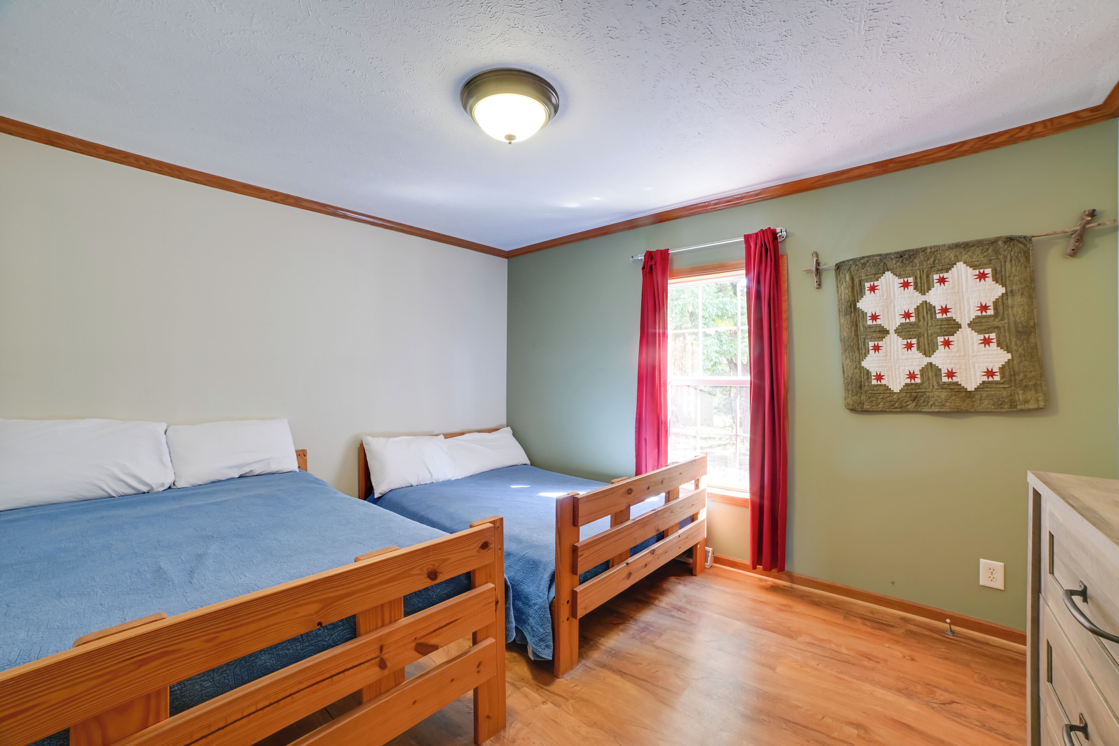 Une pièce avec deux lits en bois, une commode en bois et une courtepointe accrochée au mur.