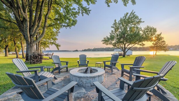 Eine Terrasse mit Adirondack-Stühlen und einer Feuerstelle aus Stein mit Blick auf einen See.