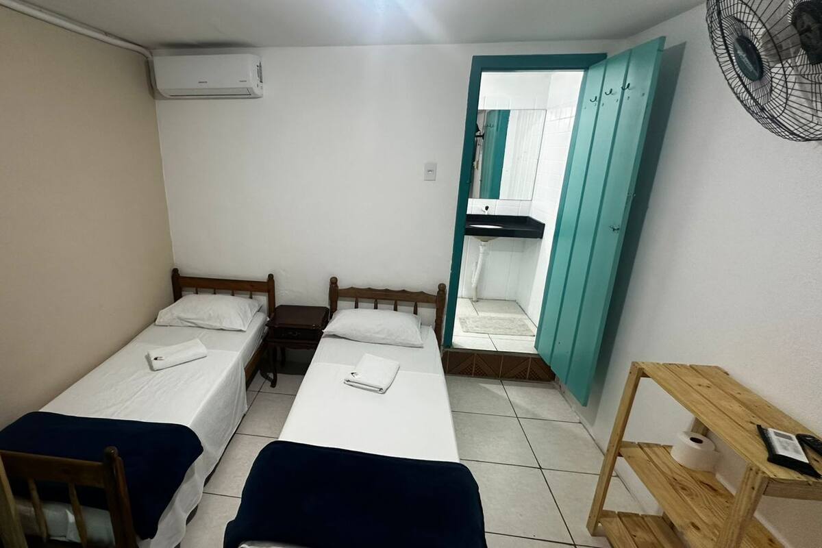 Una habitación pequeña y limpia con dos camas individuales, una mesa de madera, una silla, un ventilador y un baño visible a través de una puerta abierta.