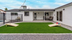 A backyard with artificial grass, two wooden play structures, a covered patio area with a table and chairs, and a white building with a grey roof. - Modern 4BR Home • Pool • South Scottsdale (Scottsdale)
