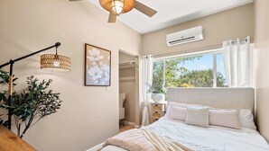 A bedroom with a bed, bedside table, lamp, air conditioning unit, window with curtains, and a view of greenery outside.