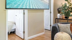 A room with a large flat-screen TV mounted on a wall, a door leading to another room, a small round table with a woven centerpiece, and a potted plant on a side table.