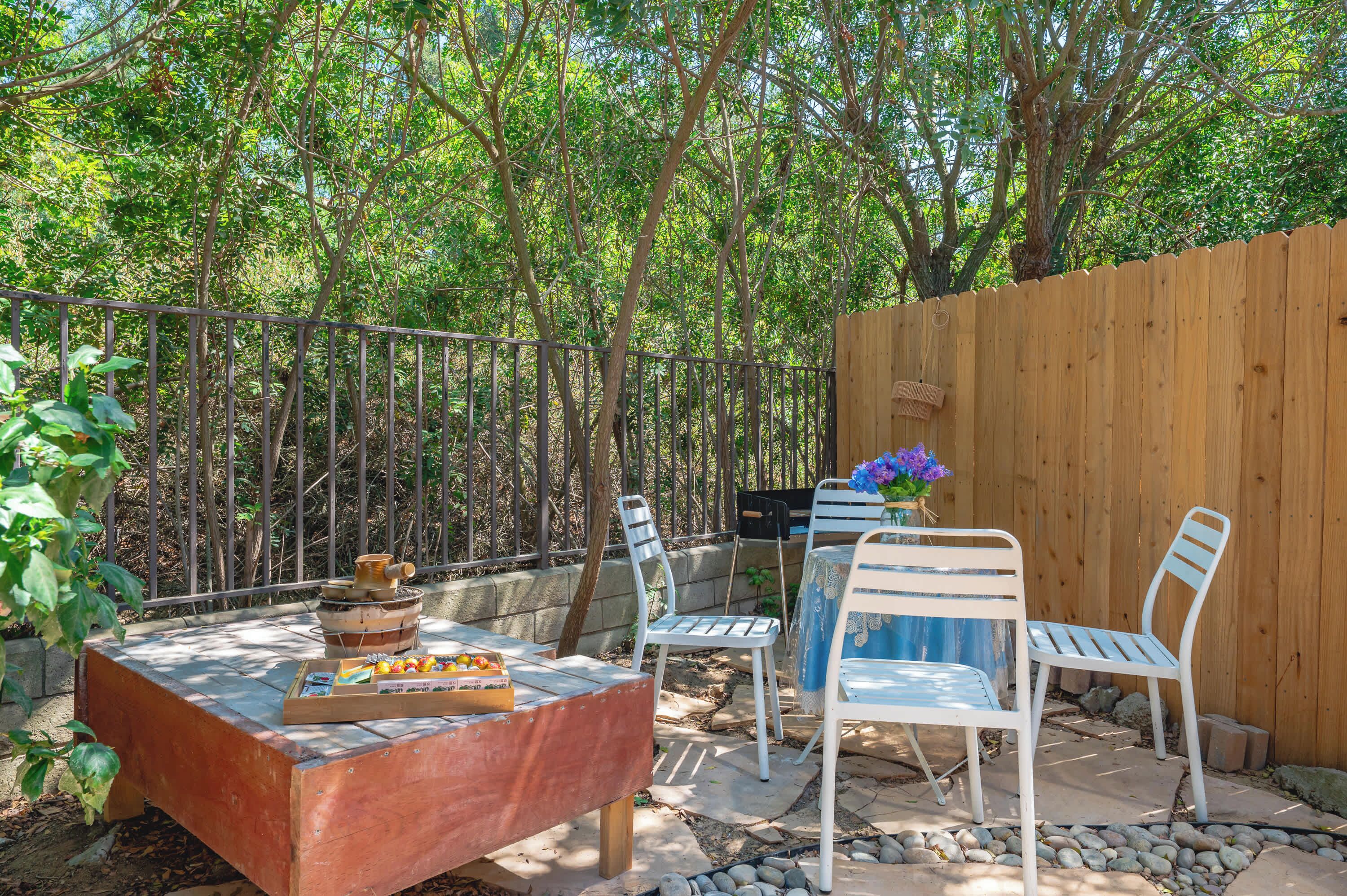 An outdoor patio with a wooden table, two white chairs, and a small garden area with a wooden fence and trees.