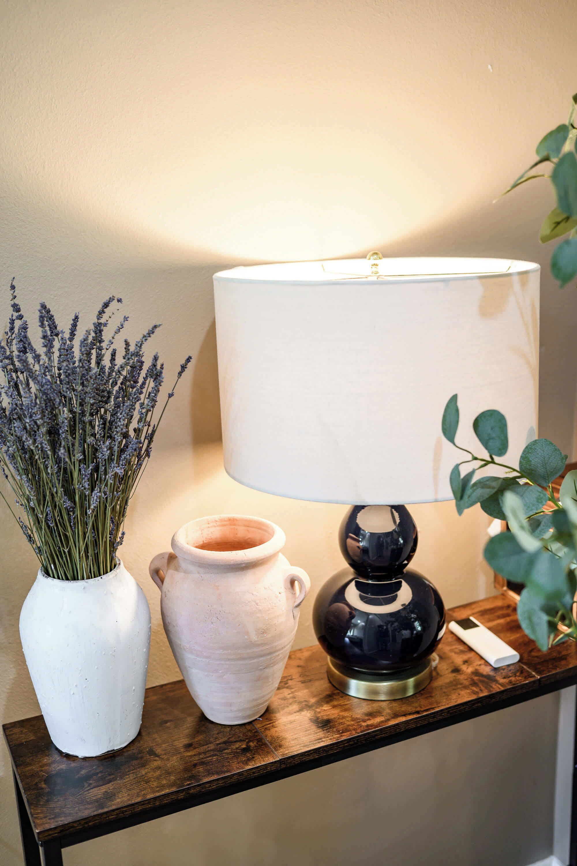A table with a white vase of lavender, a white ceramic pot, and a black lamp with a white shade.