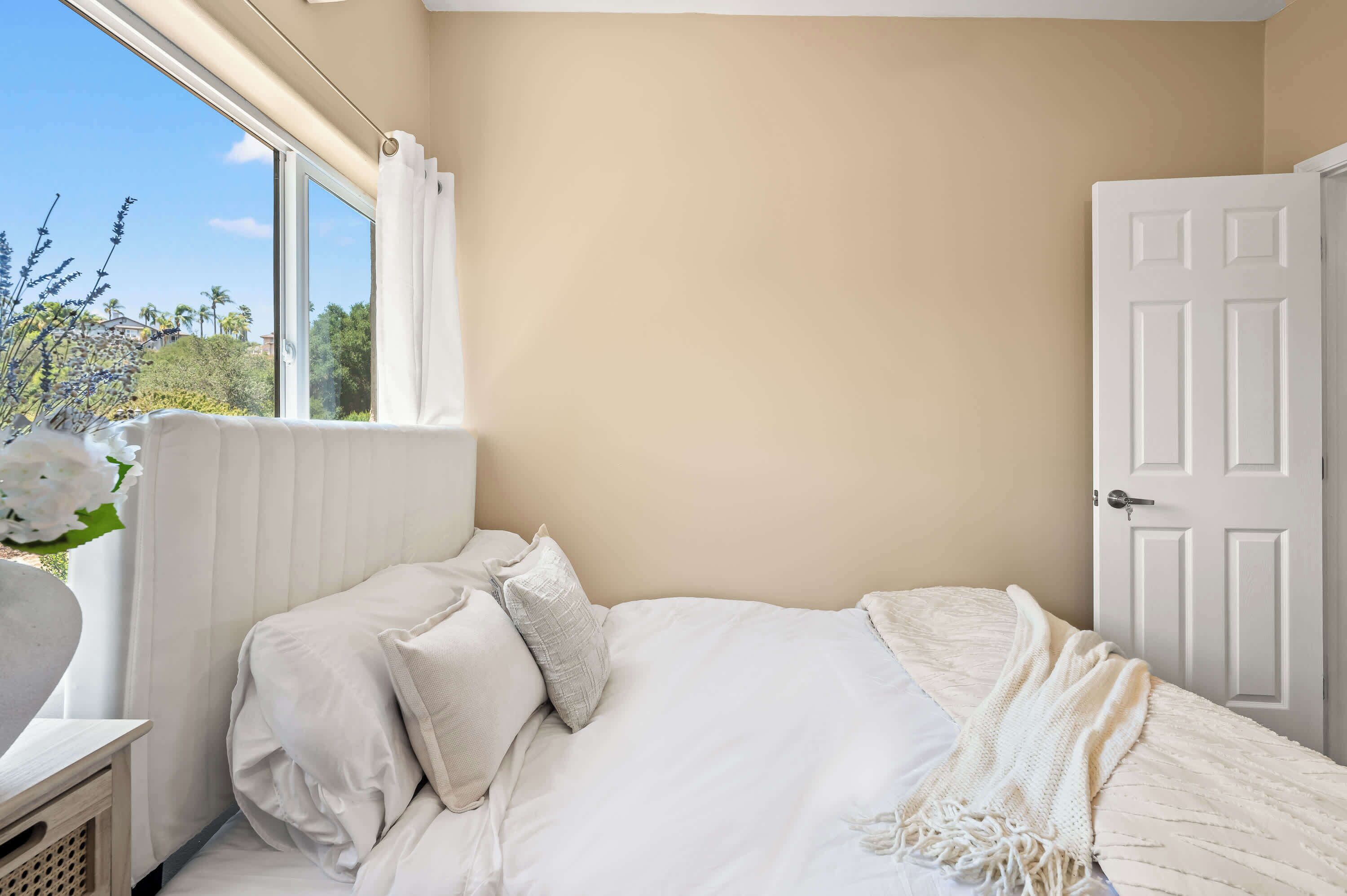 A bedroom with a bed, a window with curtains, a door, and a view of greenery outside.
